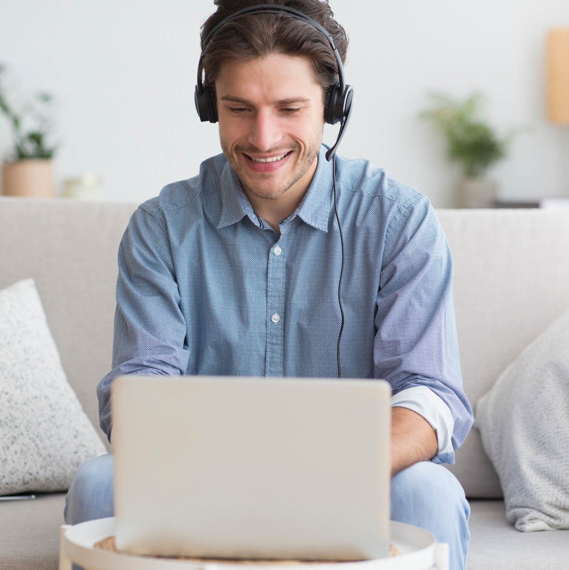 Young man sitting on a sofa with headphones smiling at his laptop screen